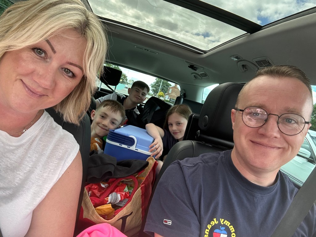 A family of five in a car, smiling and ready for a road trip, with bags and food in the back seat.