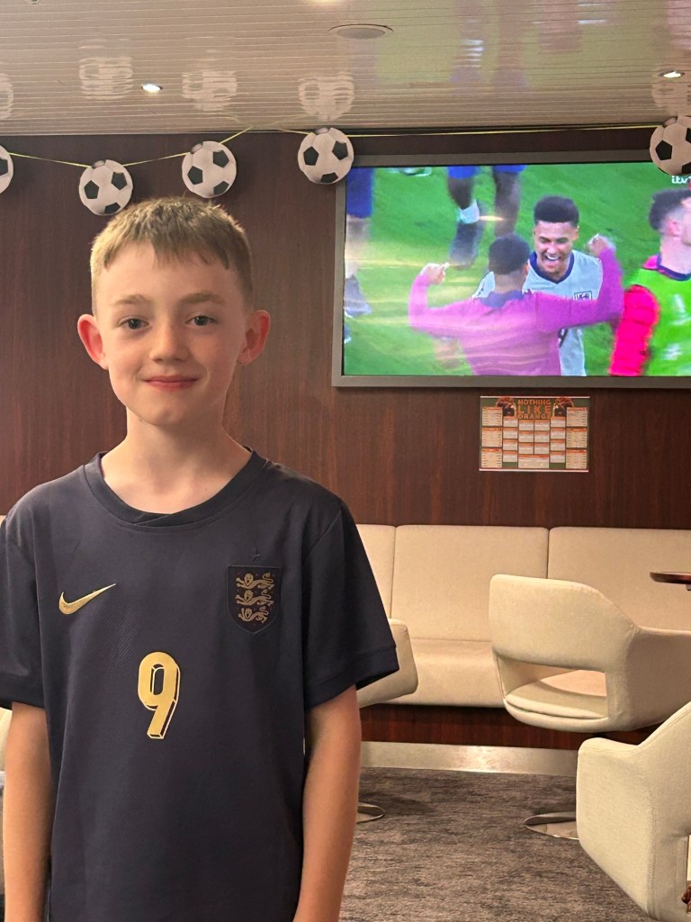 A young boy wearing an England football shirt poses in a lounge area, with a large screen displaying a football match in the background and soccer-themed decorations overhead.