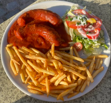 A plate of food featuring two currywurst sausages served alongside a portion of golden french fries and a fresh salad with tomatoes and lettuce.