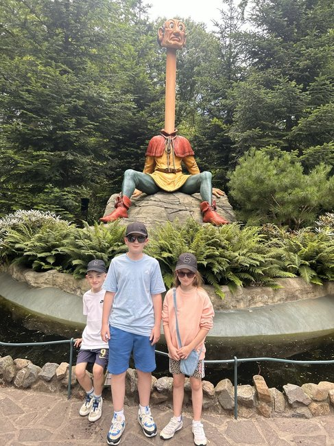 Three children posing in front of a large whimsical statue in a park, featuring a character with an elongated neck and dressed in colorful attire, surrounded by greenery.