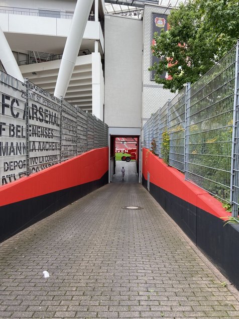 A pathway leading into a football stadium, framed by fences with team logos and banners, creating an atmosphere of anticipation.