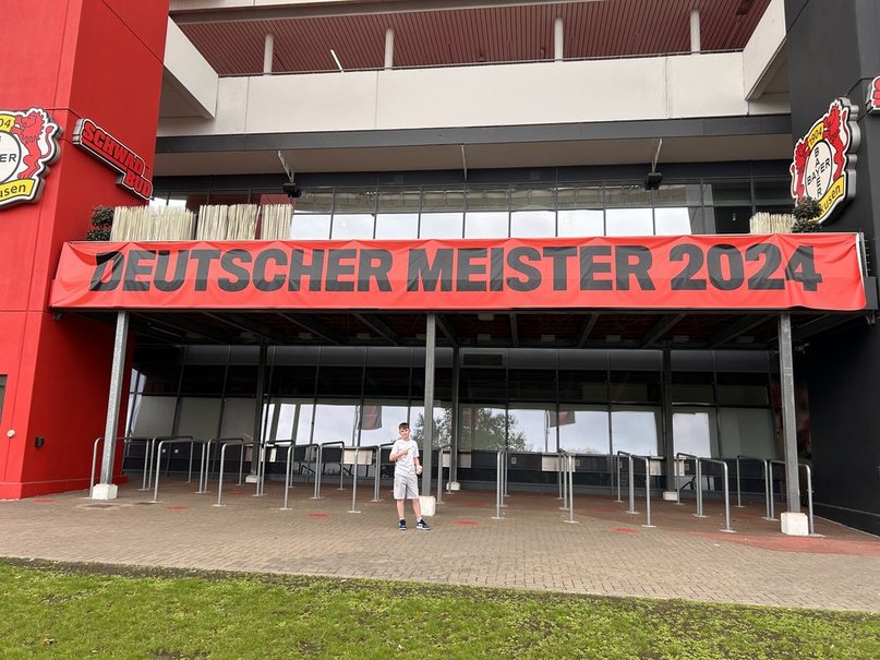 A person stands in front of Bayer Leverkusen's stadium, BayArena, featuring a large banner reading 'Deutscher Meister 2024' in bold letters.