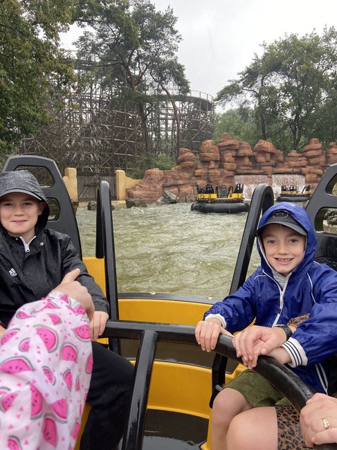Two children smiling while sitting in a water ride at a theme park, wearing raincoats. In the background, a wooden roller coaster and rocky structures are visible.