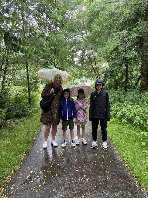 A woman holding an umbrella stands on a path with three children, surrounded by greenery on a rainy day.