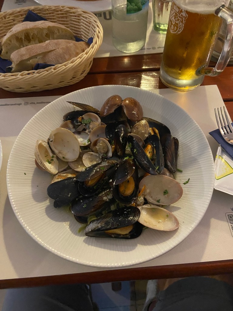 A plate of fresh seafood including mussels and clams, accompanied by a basket of bread and a glass of beer.