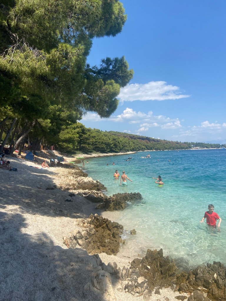 A sunny beach in Croatia with clear turquoise water, rocky shores, and people enjoying the water and relax under the trees.