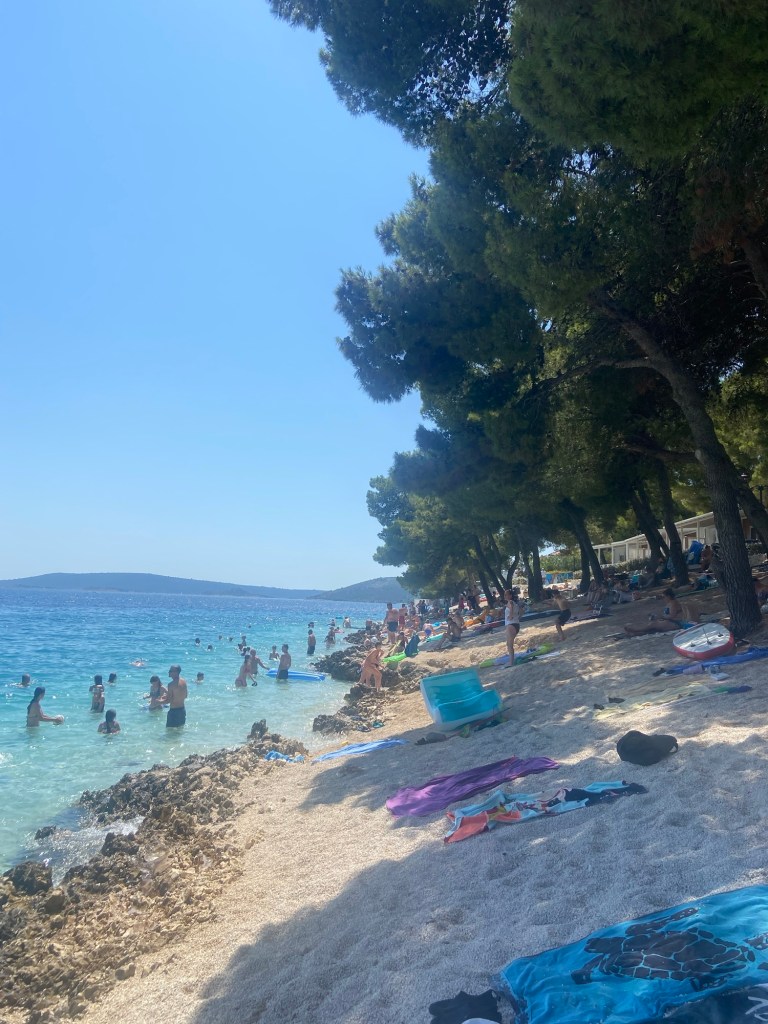 A busy beach scene in Croatia with people swimming in the clear blue water, sandy areas lined with colorful towels, and tall trees providing shade along the shore.