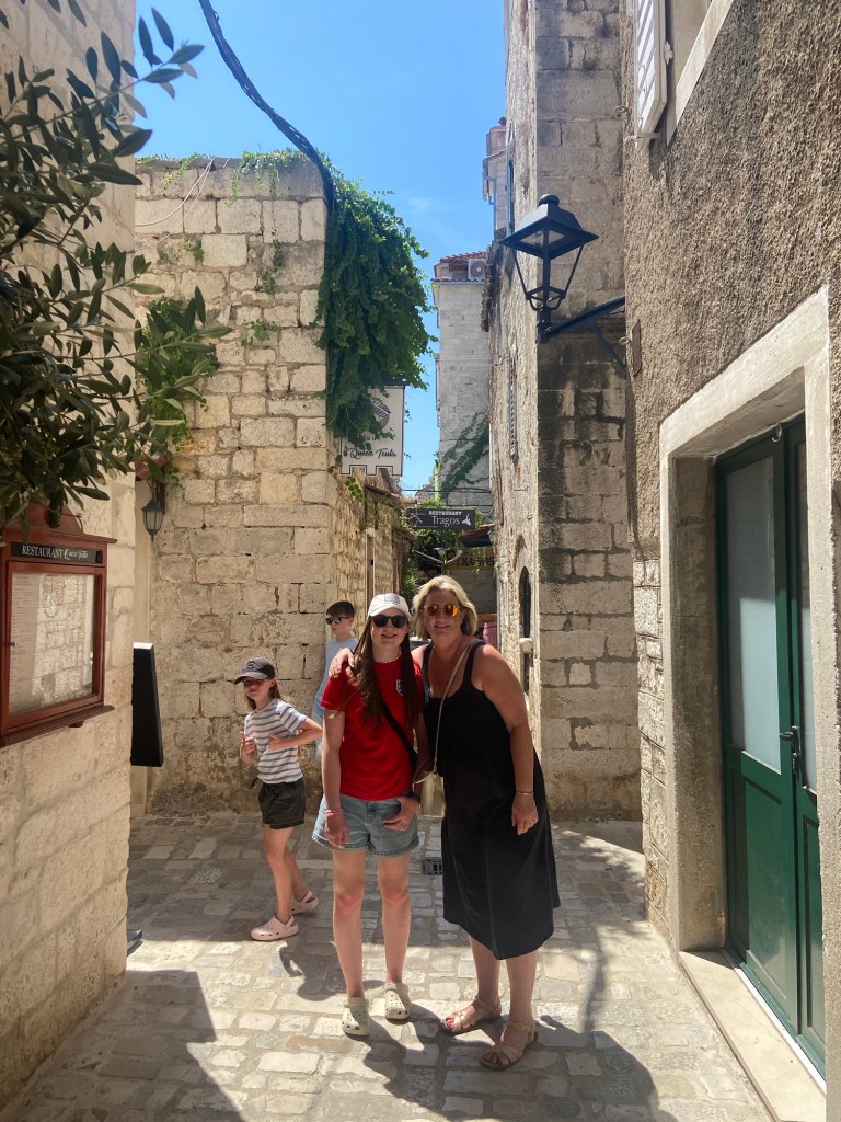 Two women pose together in a narrow, sunlit alleyway lined with stone buildings in a historic town, with greenery visible overhead. A child can be seen in the background.