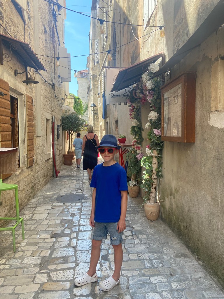 A child wearing a blue shirt and sunglasses stands on a cobblestone street in a narrow alleyway, with a backdrop of stone buildings and decorative plants.