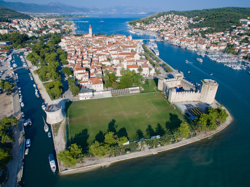 Aerial view of Trogir, Croatia, showcasing the historic town with red-roofed buildings, a waterfront marina, and ancient stone fortifications surrounded by lush greenery and the Adriatic Sea.