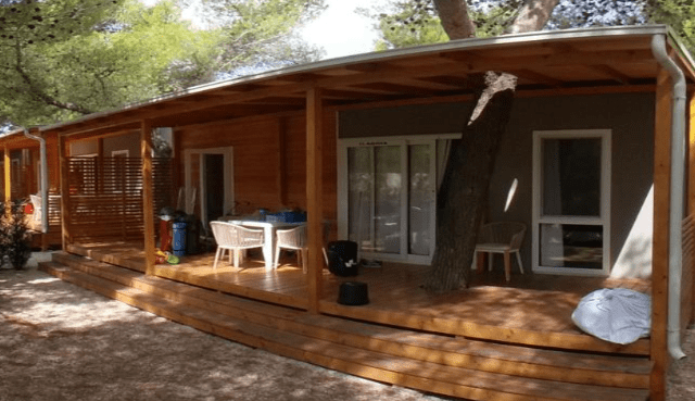 Exterior view of a wooden mobile home at a campsite in Croatia, featuring a spacious deck, outdoor seating, and a tree integrated into the structure.