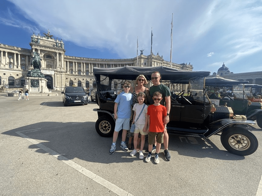 The Murtagh Family posing in front of a classic car tour vehicle in front of the Hofsburg Palace, Vienna on a sunny day